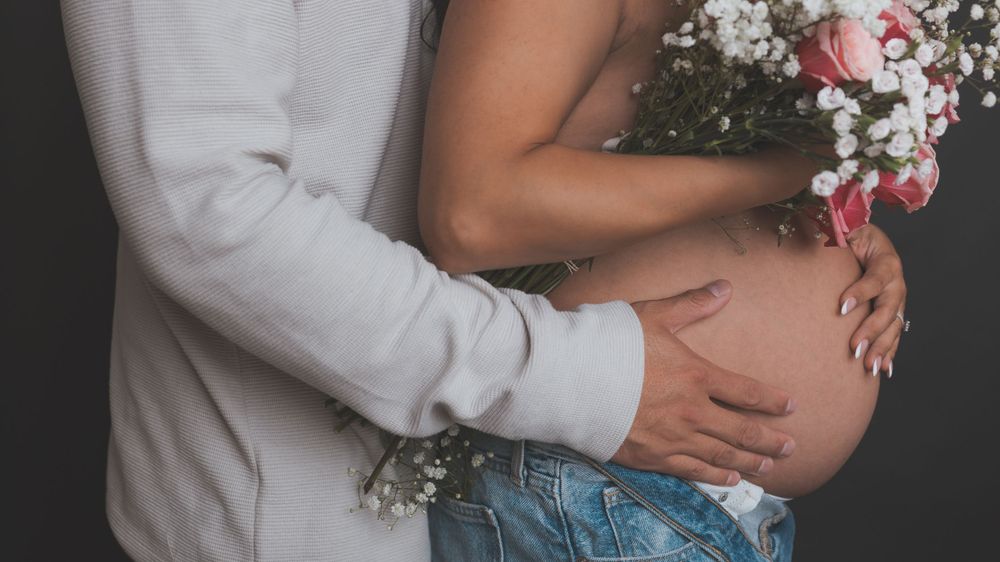 Close-up of hands embracing a pregnant belly with pink flowers in frame.