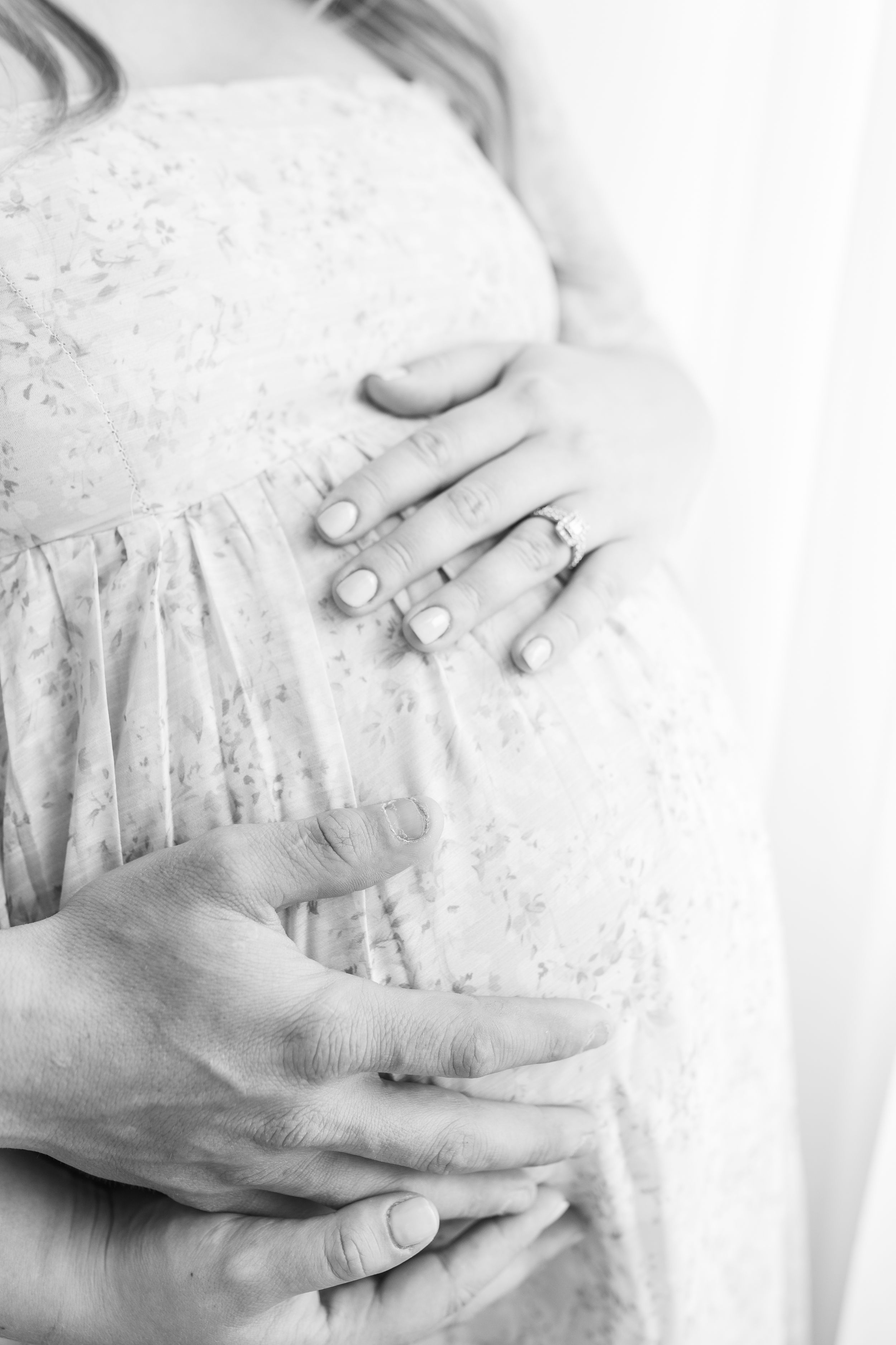 Black and white photo of a pregnant woman in floral dress with hands cradling her baby bump.
