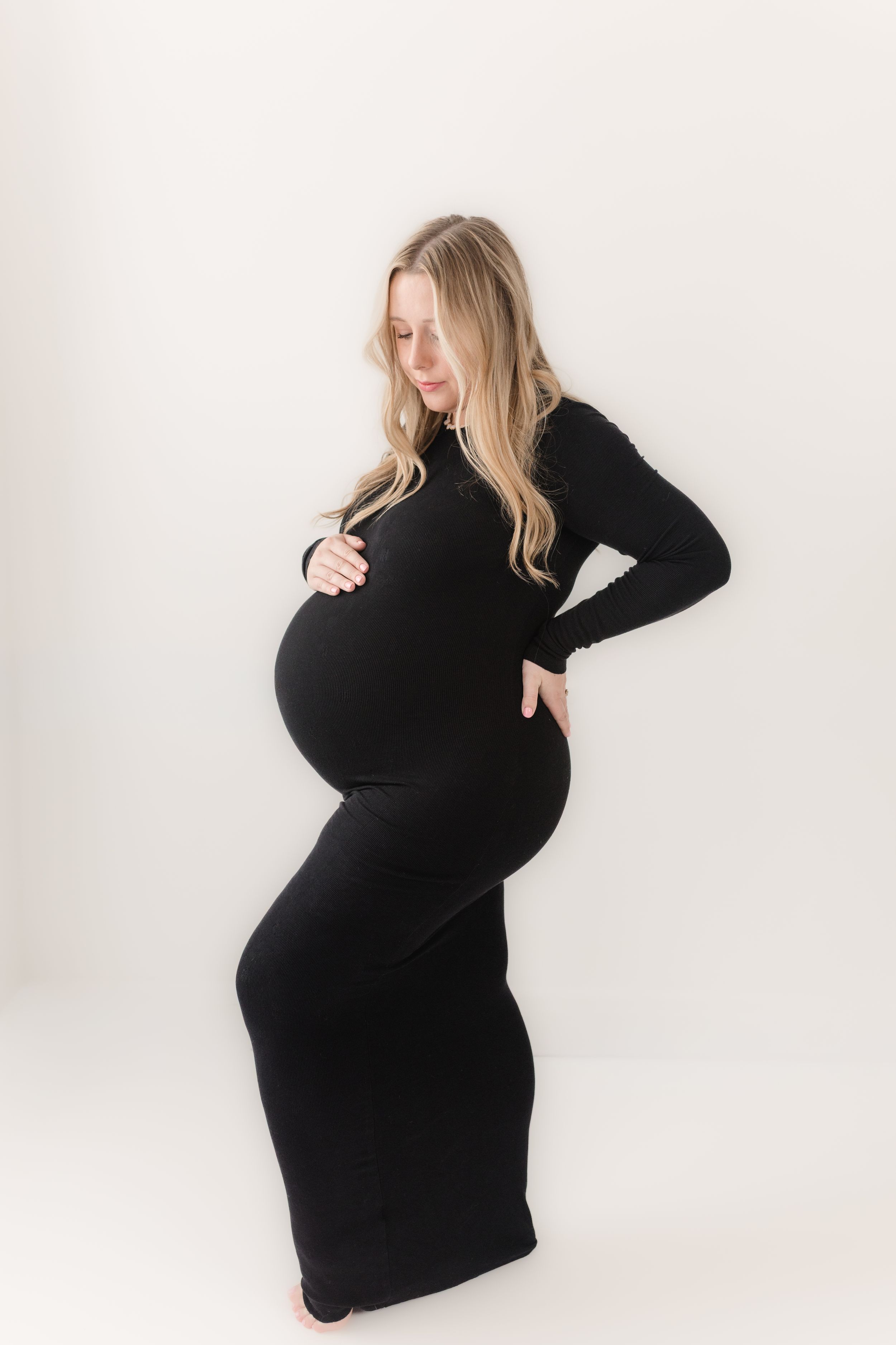 Pregnant woman in black form-fitting dress cradling baby bump against white background.