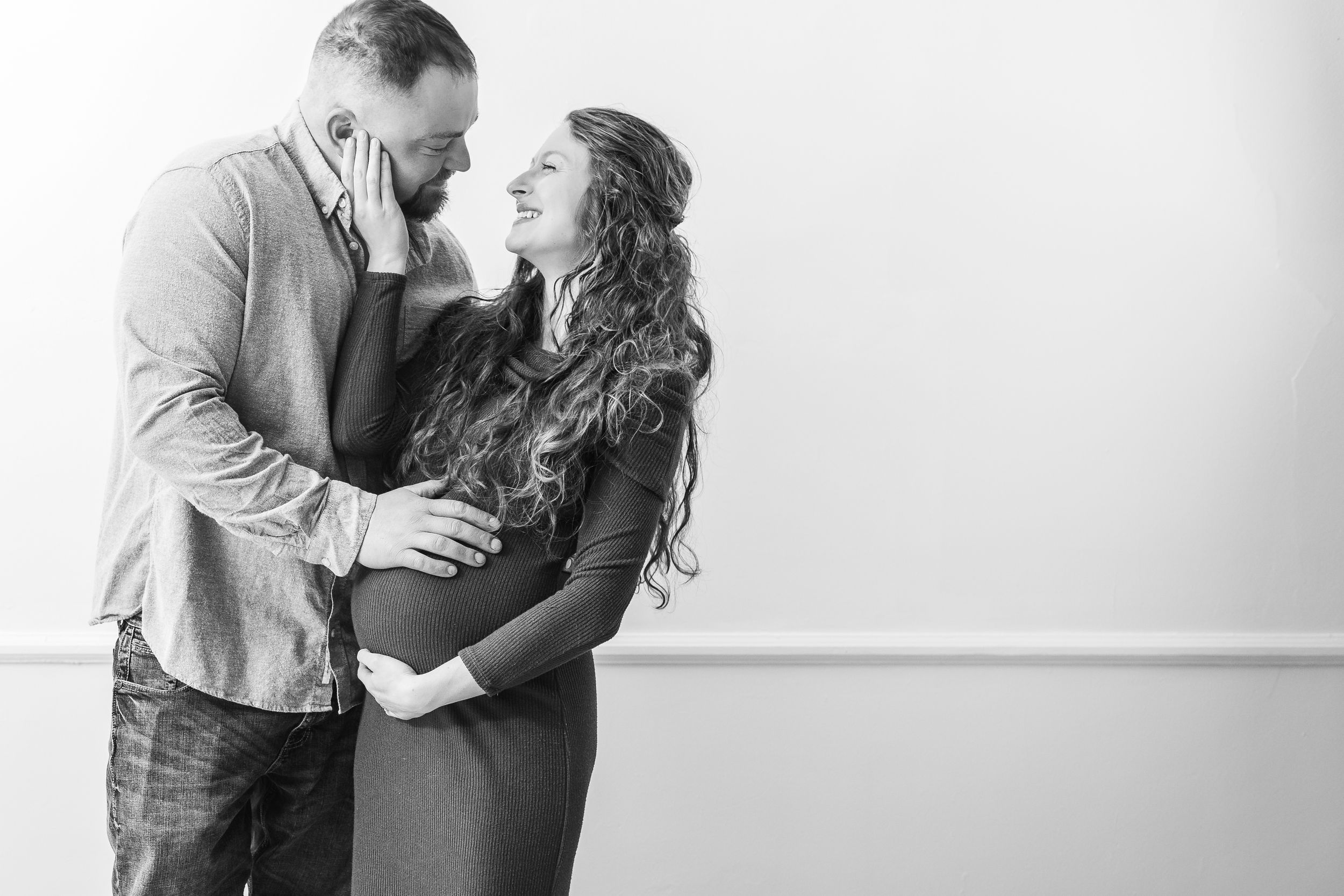 Black and white maternity photo of couple embracing against plain wall.