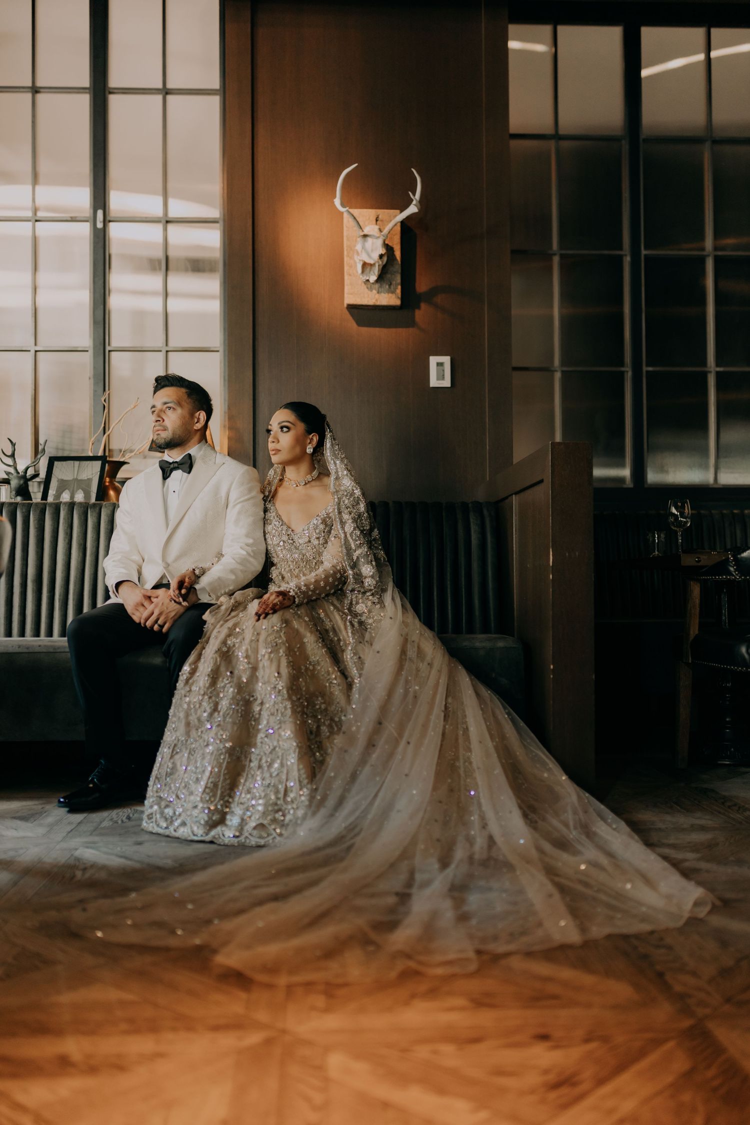 Wedding couple seated in elegant attire against dark wall with mounted antlers decor.