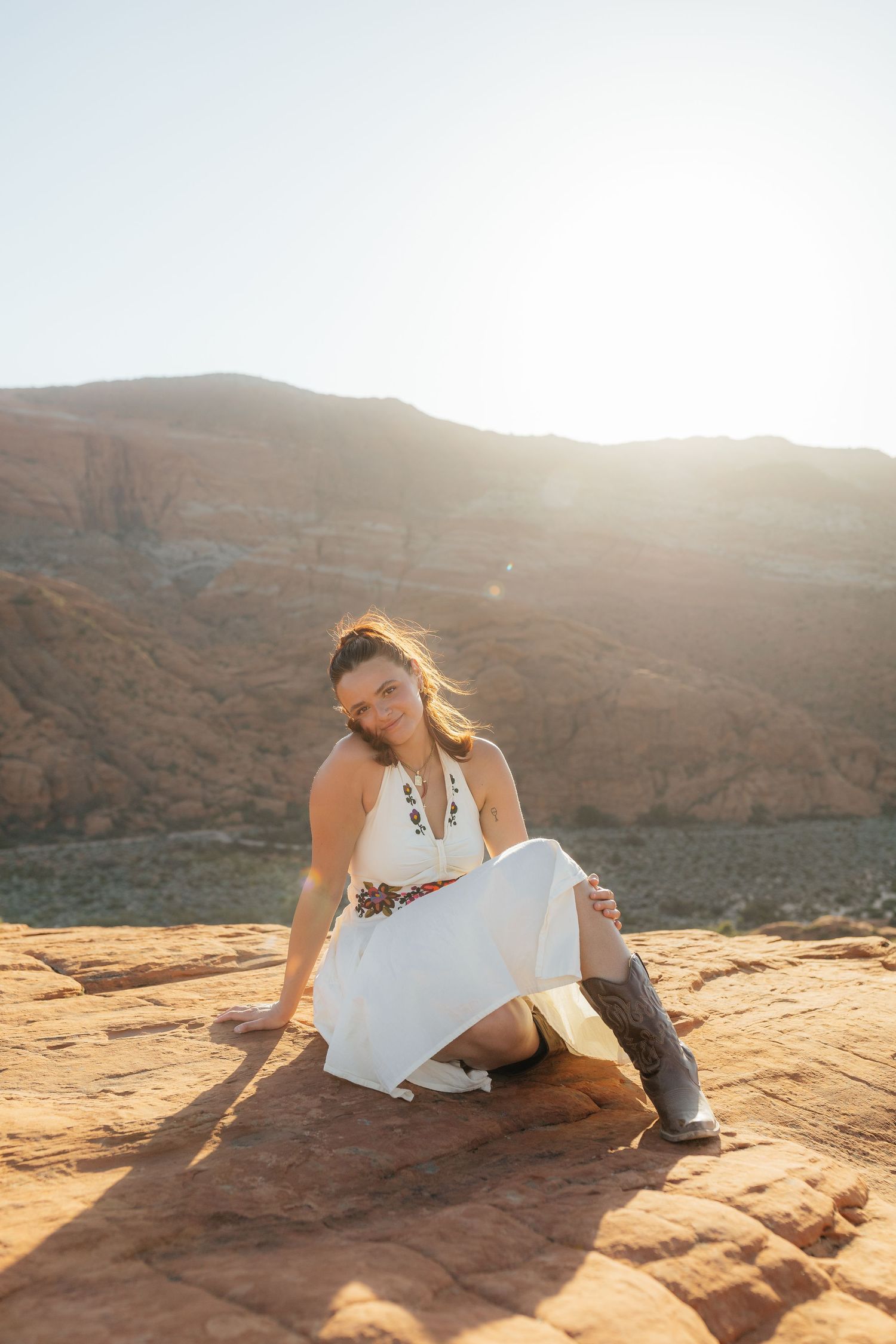Woman in a white halter dress and cowboy boots sits on red rock terrain with sunlit desert mountains behind her.