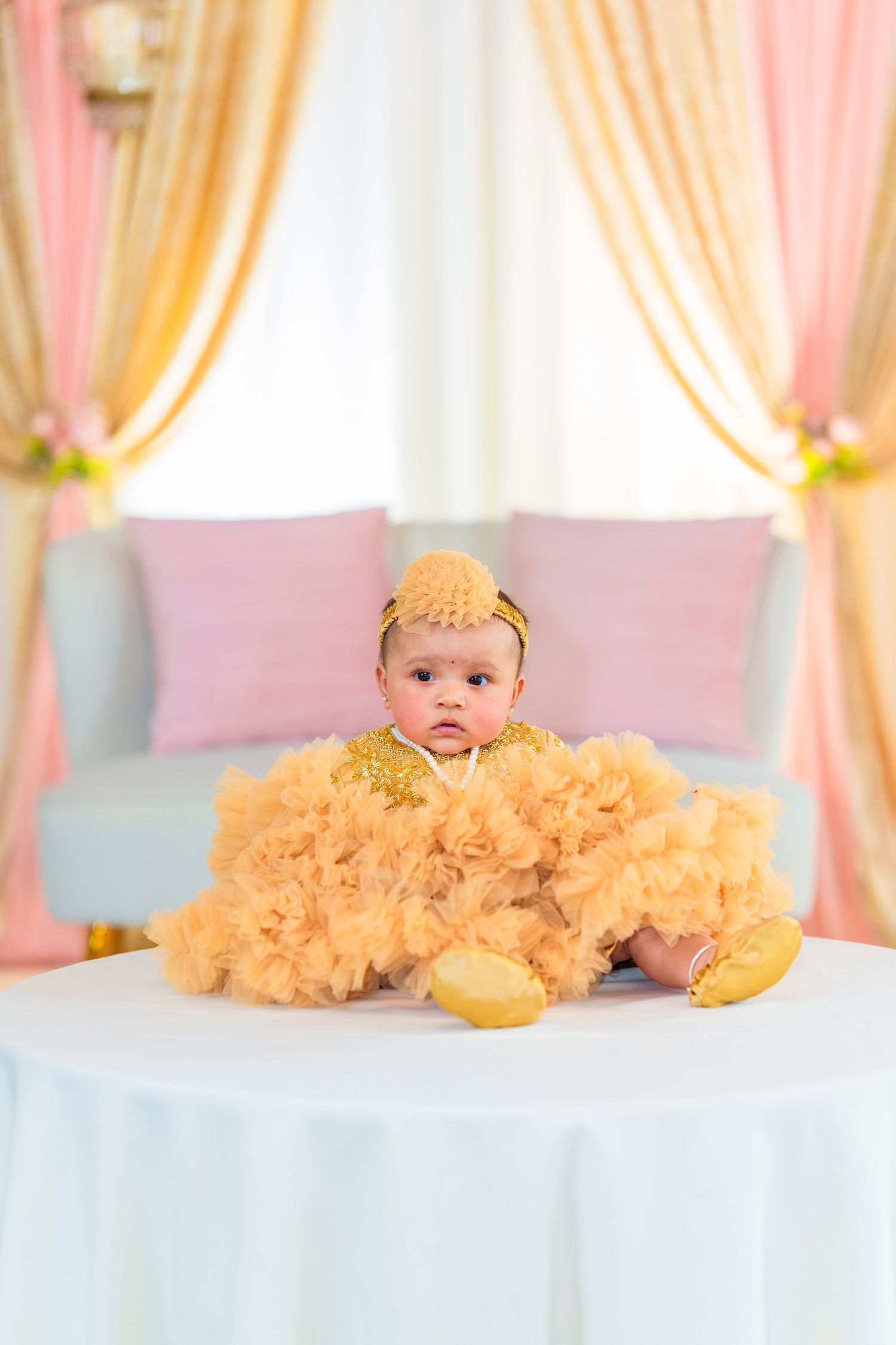 A baby wearing a yellow tulle dress and headband sits on a couch with pink and gold curtains in the background.