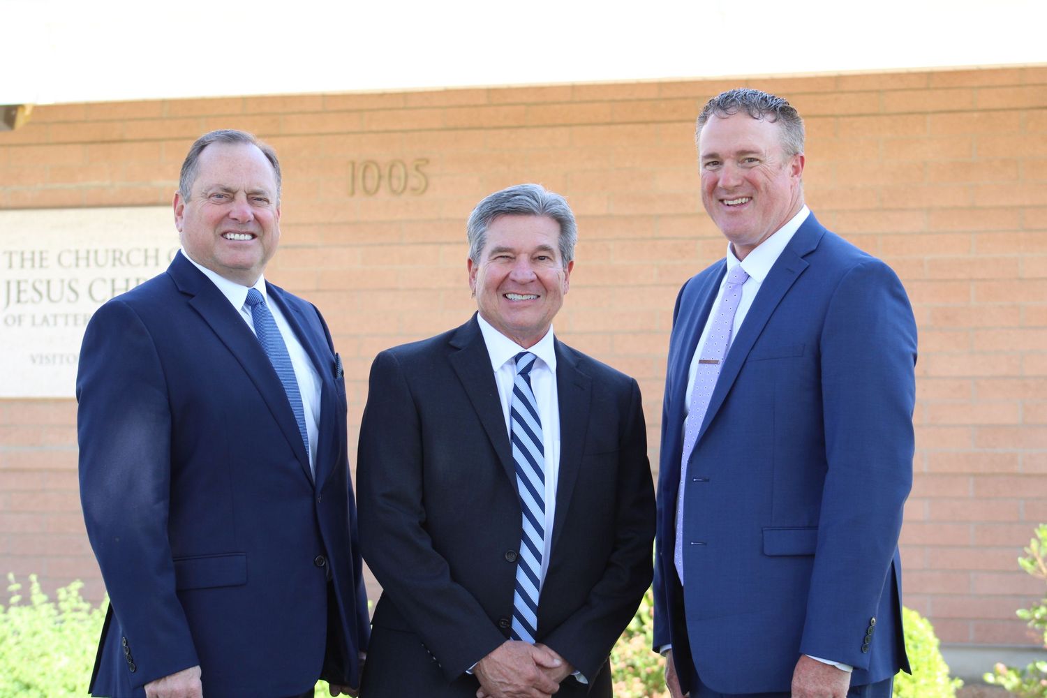 Three businessmen in navy suits pose for a photo outside an office building.