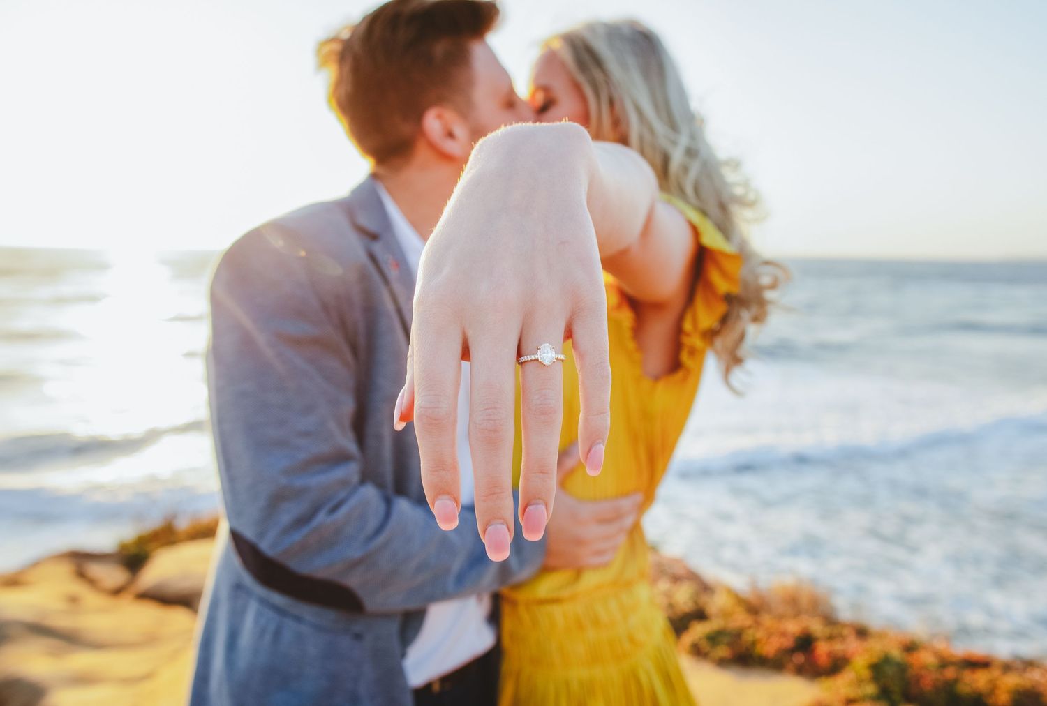 man proposing to woman and her showing off engagement ring at sunset cliffs