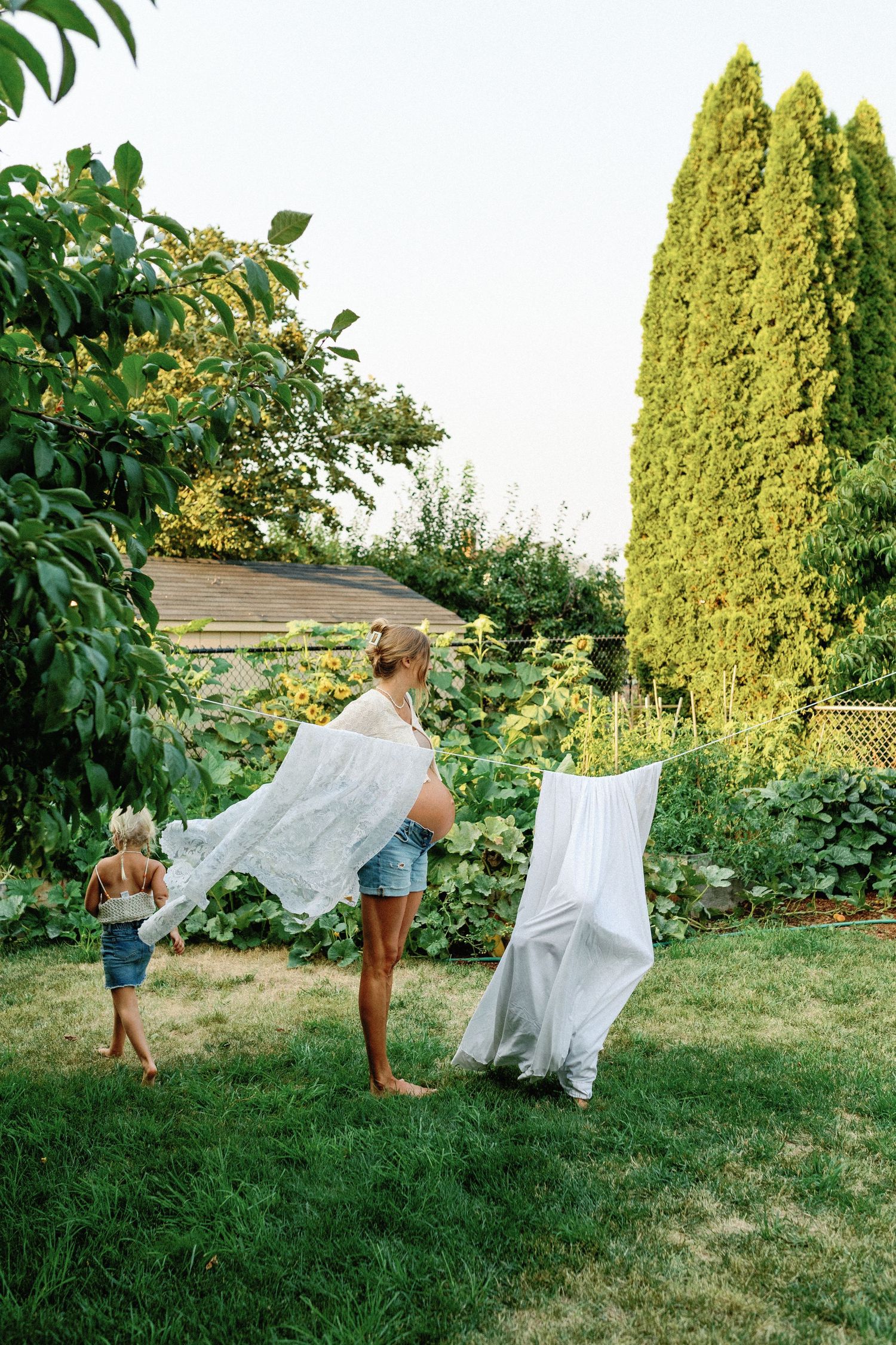 People hanging white linens on a clothesline in a lush backyard garden with tall cypress trees on a sunny day.