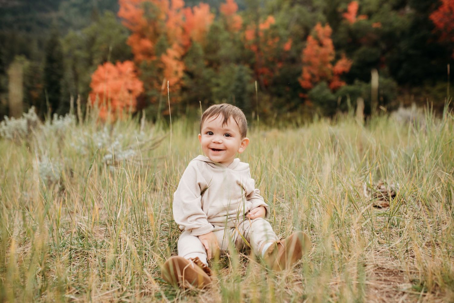 A small child sits in autumn grass wearing neutral clothing with fall foliage behind.
