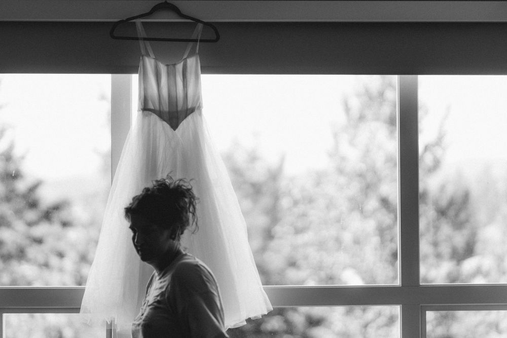 A silhouetted figure stands before a hanging wedding dress against a bright window.