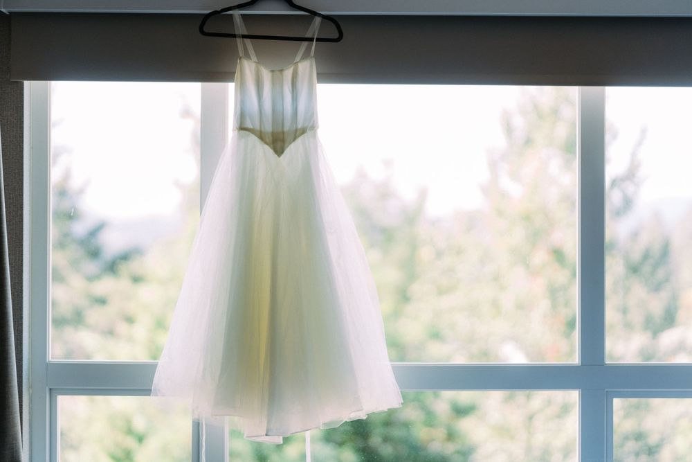 White wedding dress hanging near a sunlit window for a pre-ceremony photo.