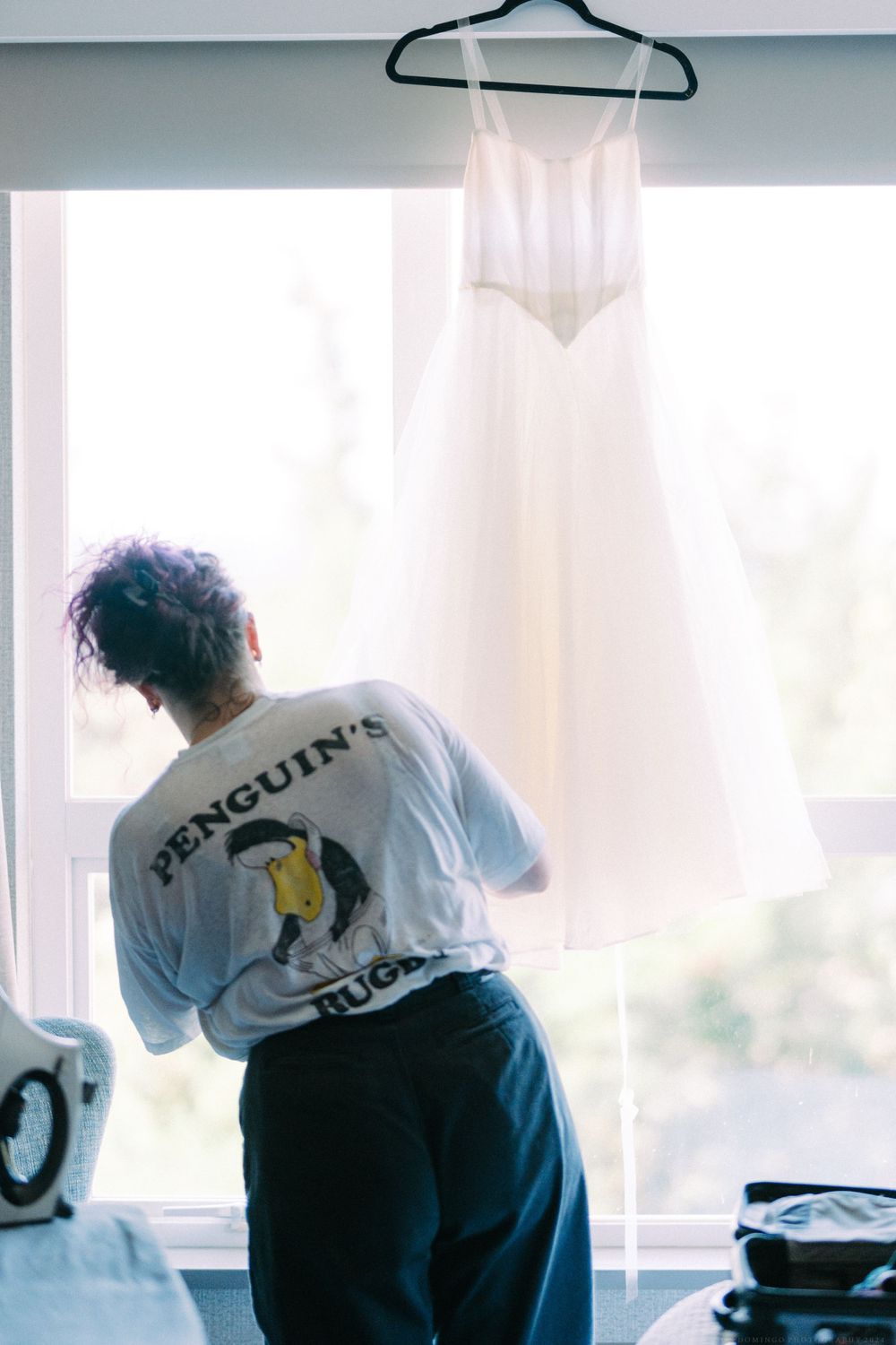 Someone in a penguin t-shirt stands near a hanging white wedding dress by a bright window.