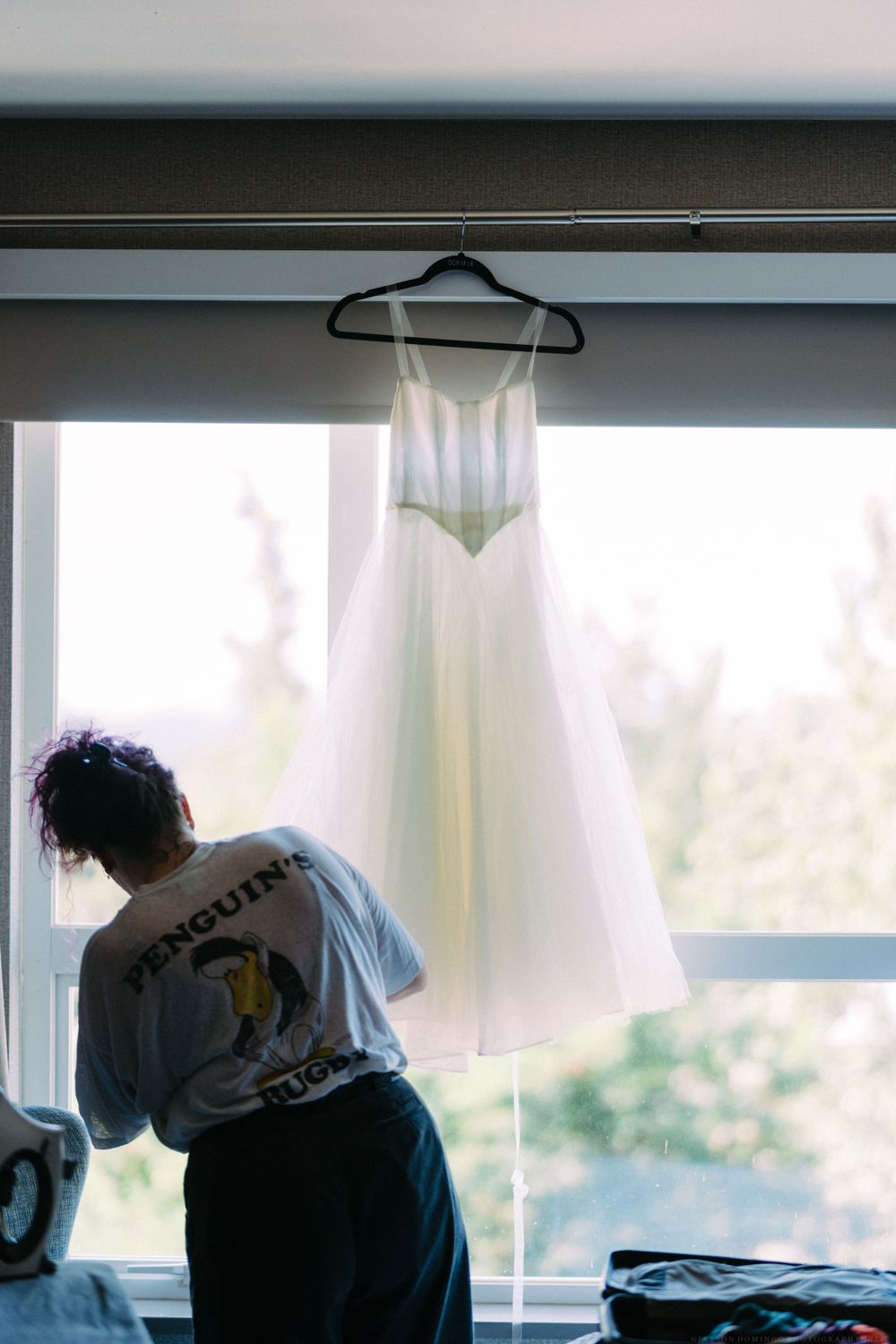 A white wedding dress hangs by a window while someone adjusts it.