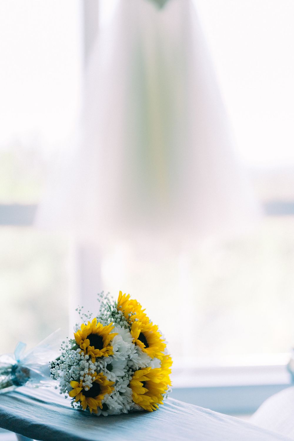 A sunflower and baby's breath bouquet sits near a white dress by a window.