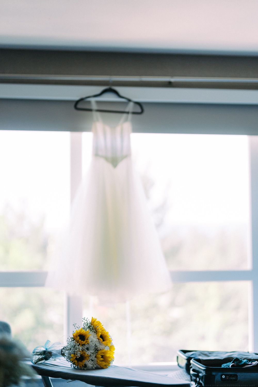 Elegant white wedding dress suspended by a hanger with yellow sunflowers bouquet below.