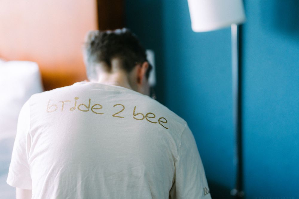 Back view of white t-shirt with 'bride 2 bee' text against blue wall.