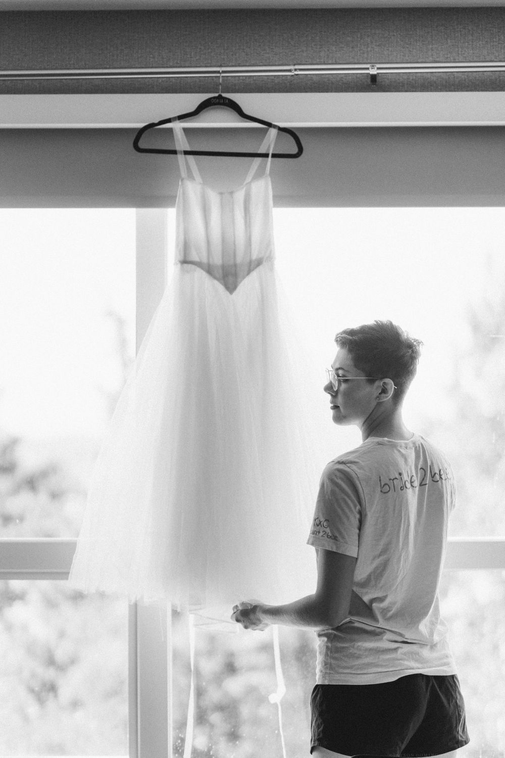 A elegant white wedding dress hangs by a window with someone standing nearby.