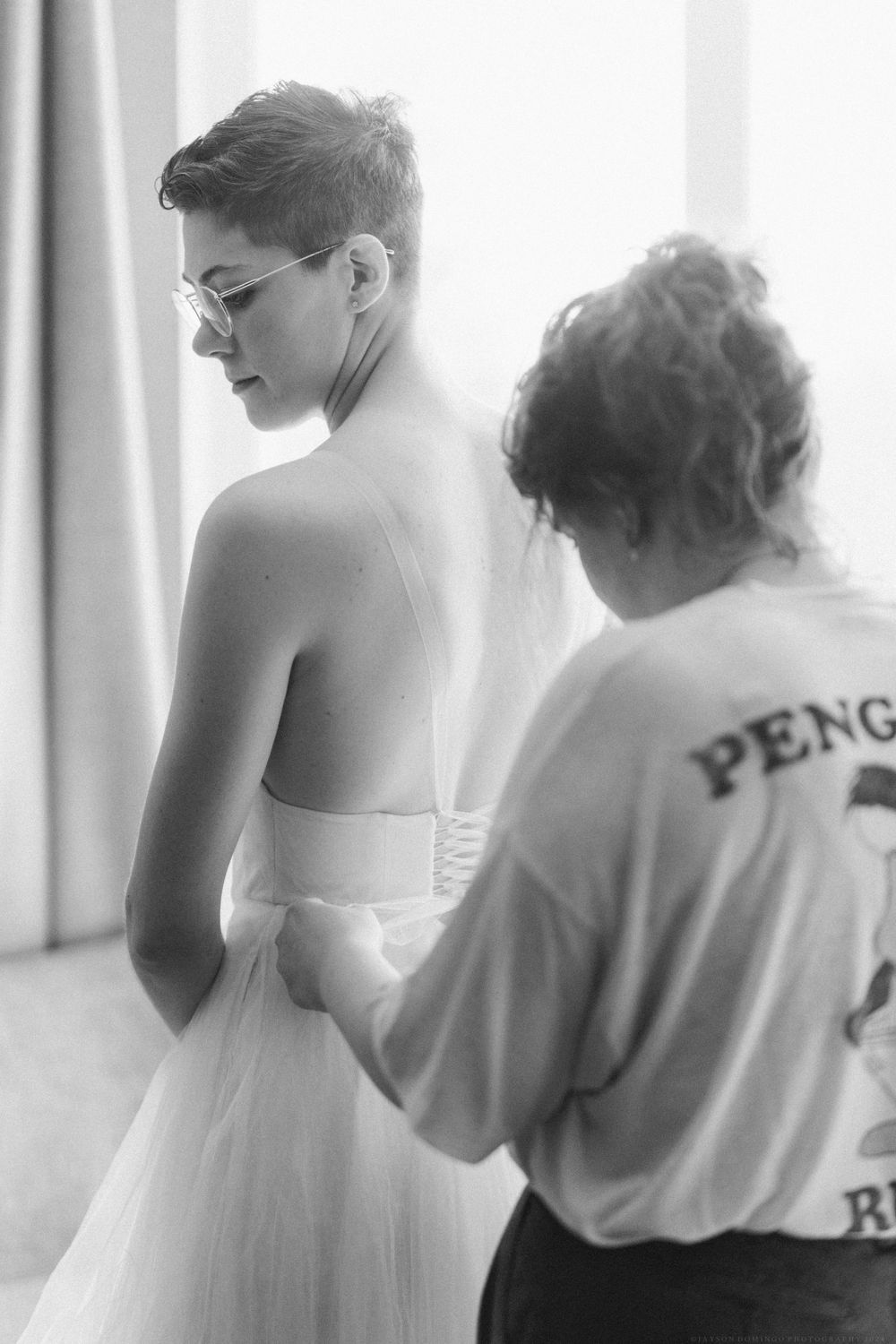 Black and white image showing a wedding dress fitting with helper in penguin shirt.
