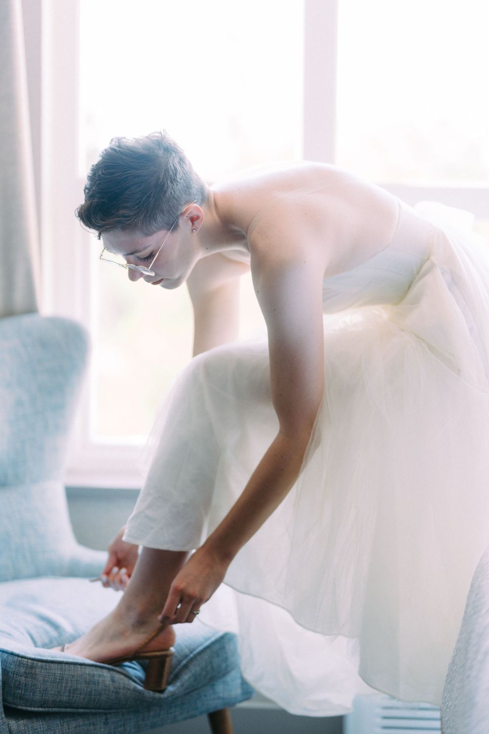 Person in white wedding dress bending down to adjust shoes by a window.