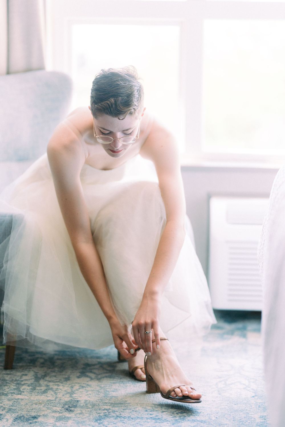Person in white wedding dress sitting while fastening sandals on blue carpet.