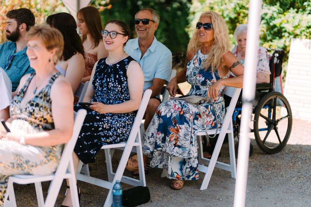 Wedding guests seated in white folding chairs at an outdoor ceremony on a sunny day.