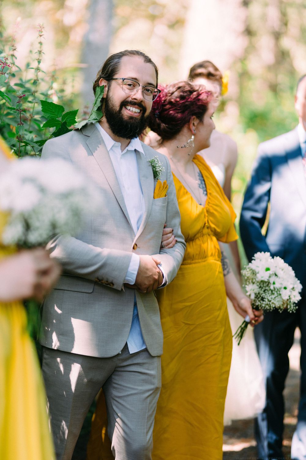 Wedding party members in formal attire including a grey suit and yellow dress standing among greenery.