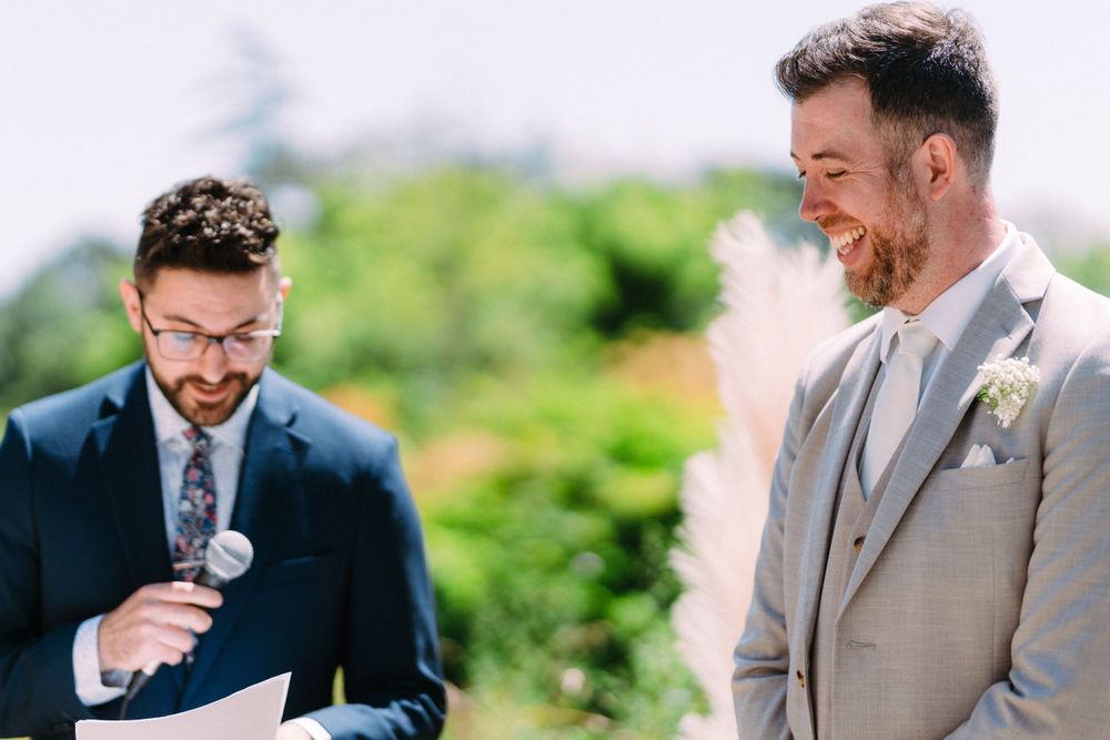 Two wedding guests in suits share a lighthearted moment during an outdoor ceremony.