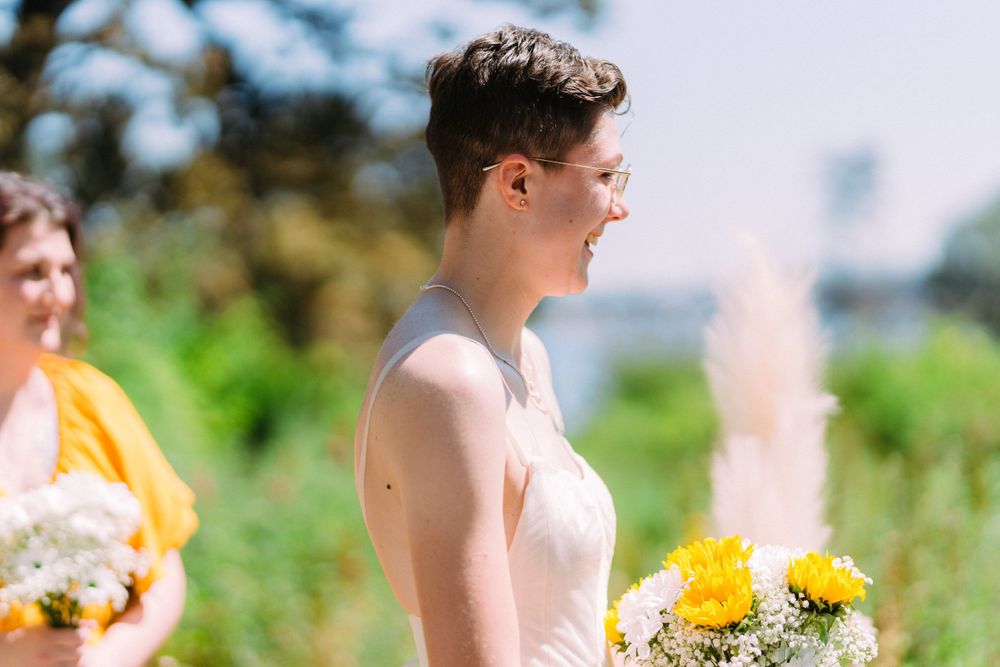 A person in a white wedding dress holds a yellow and white bouquet during a sunny outdoor ceremony.