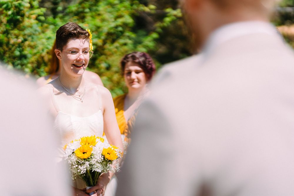 A smiling person in a wedding dress holds yellow flowers while standing outdoors.