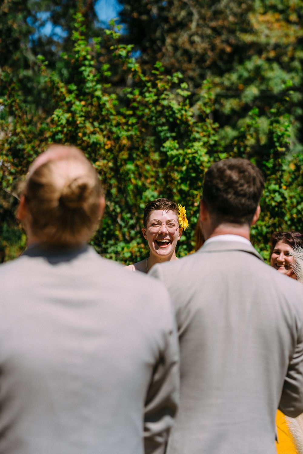 Wedding guests in light colored suits watch joyfully during an outdoor ceremony.