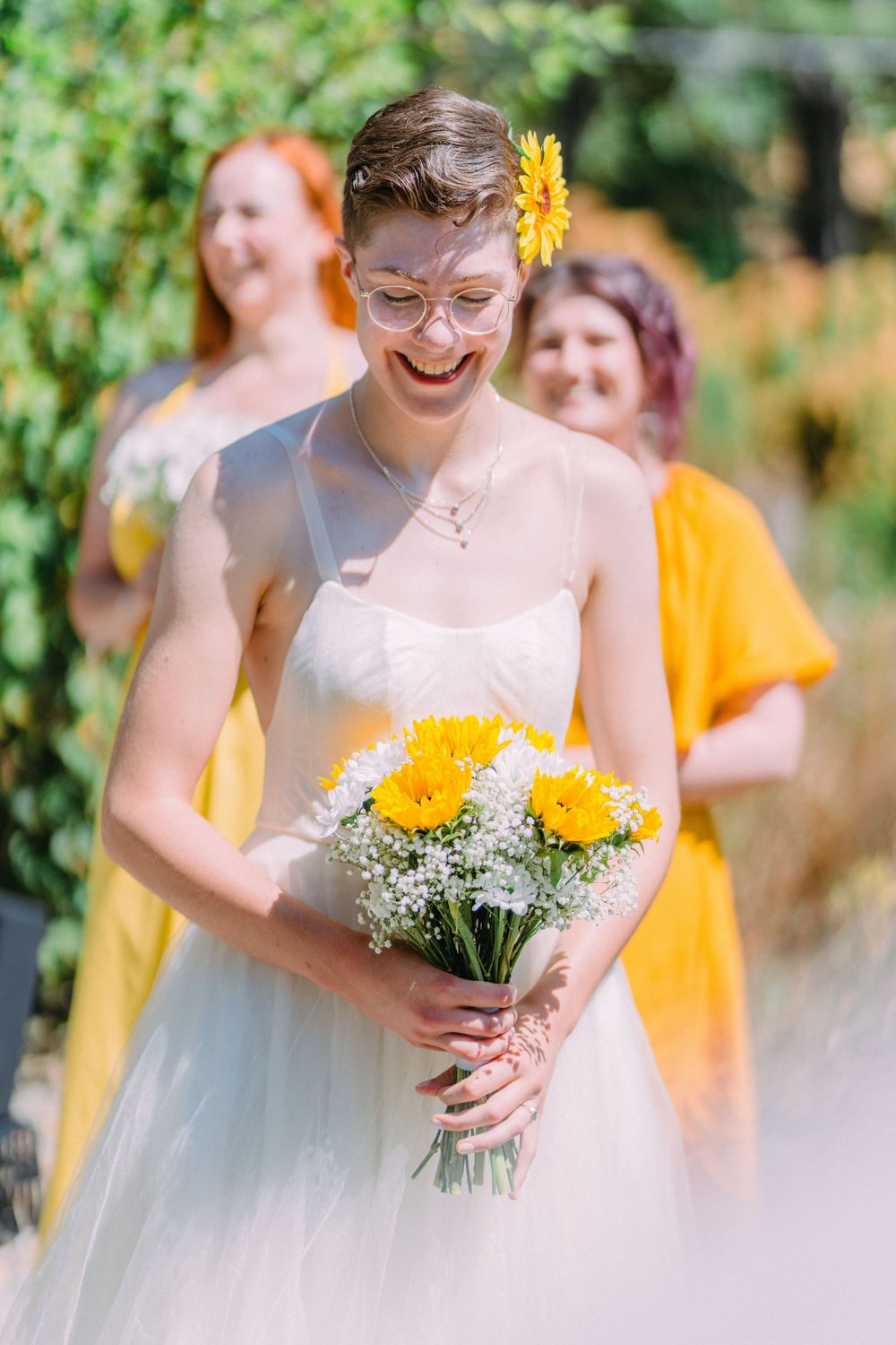 A person in a white wedding dress holds yellow flowers while laughing outdoors.