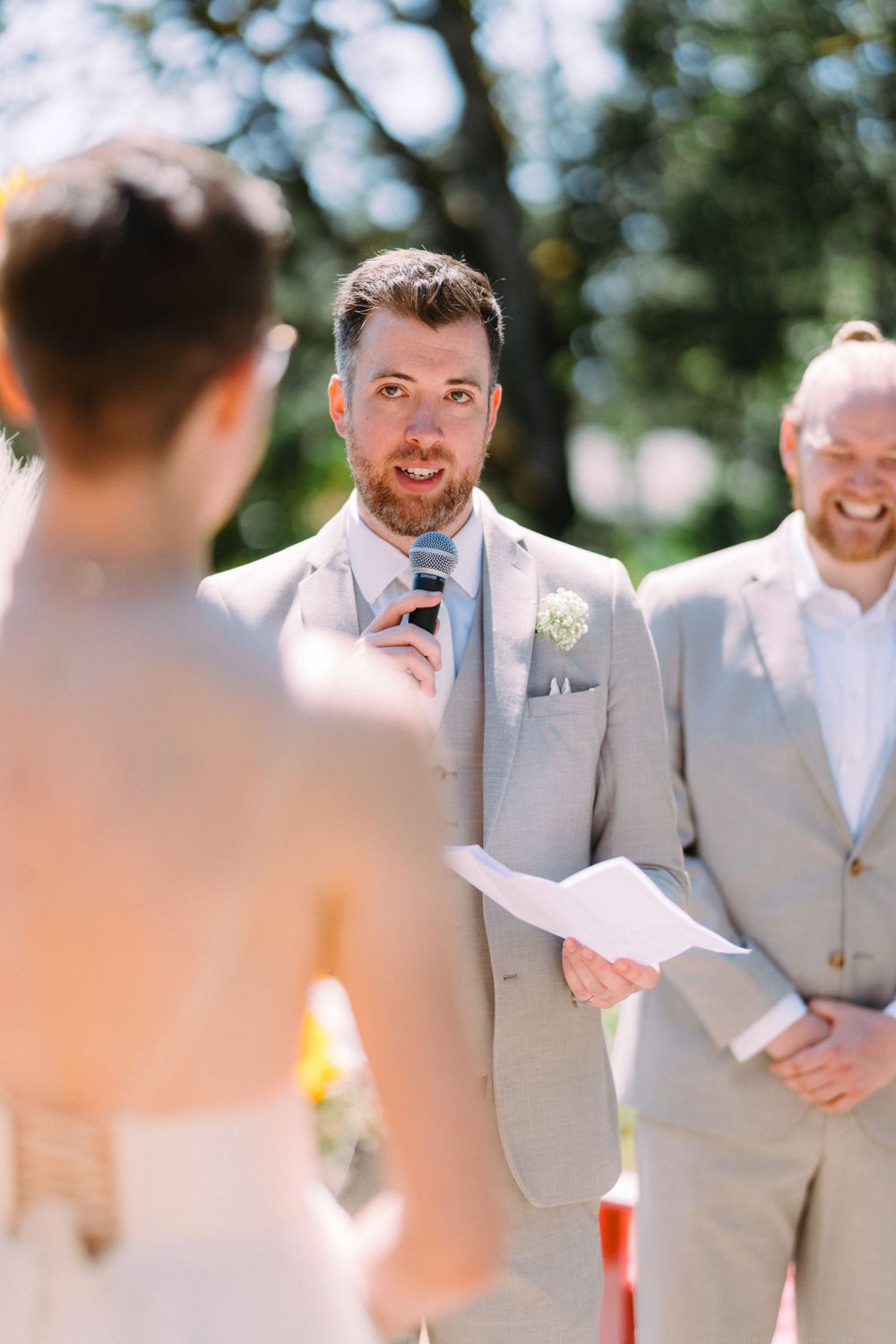 A person in a light gray suit speaks into a microphone during an outdoor wedding ceremony.