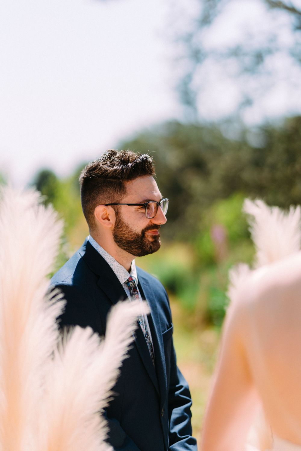 Groom in navy suit and glasses stands amid decorative pampas grass at outdoor wedding.