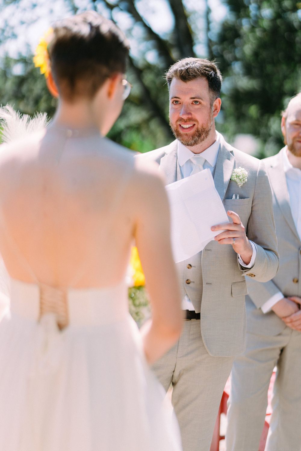 Groom in light gray suit smiles while reading vows during outdoor wedding ceremony.