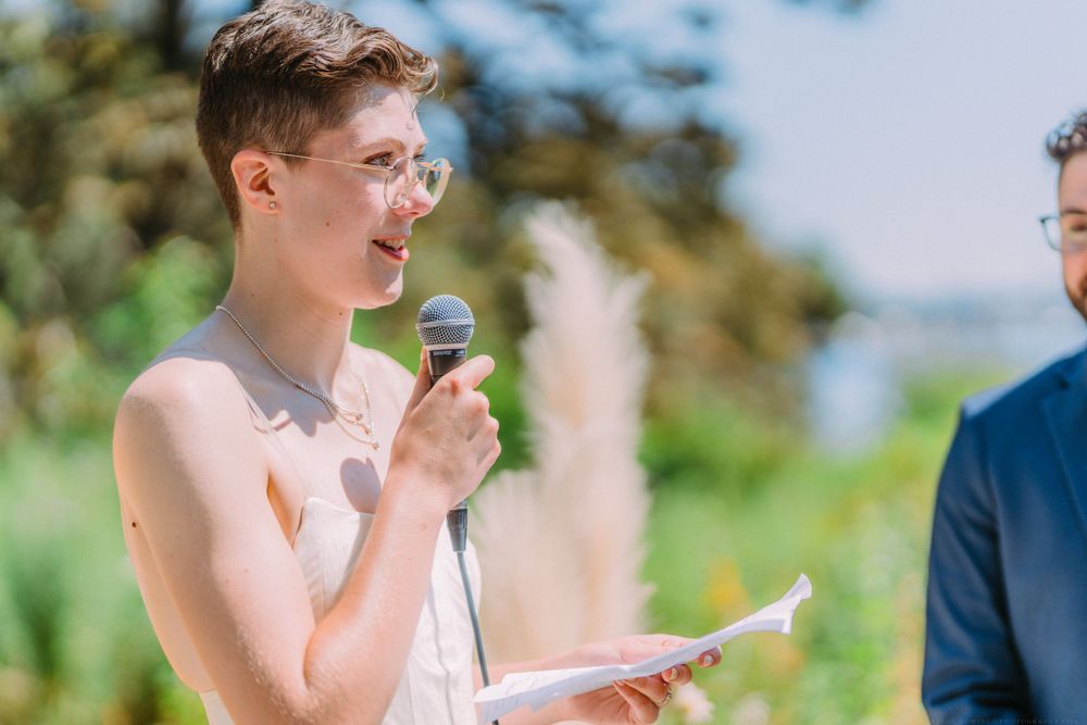 Wedding ceremony participant speaks into microphone against sunny outdoor backdrop.