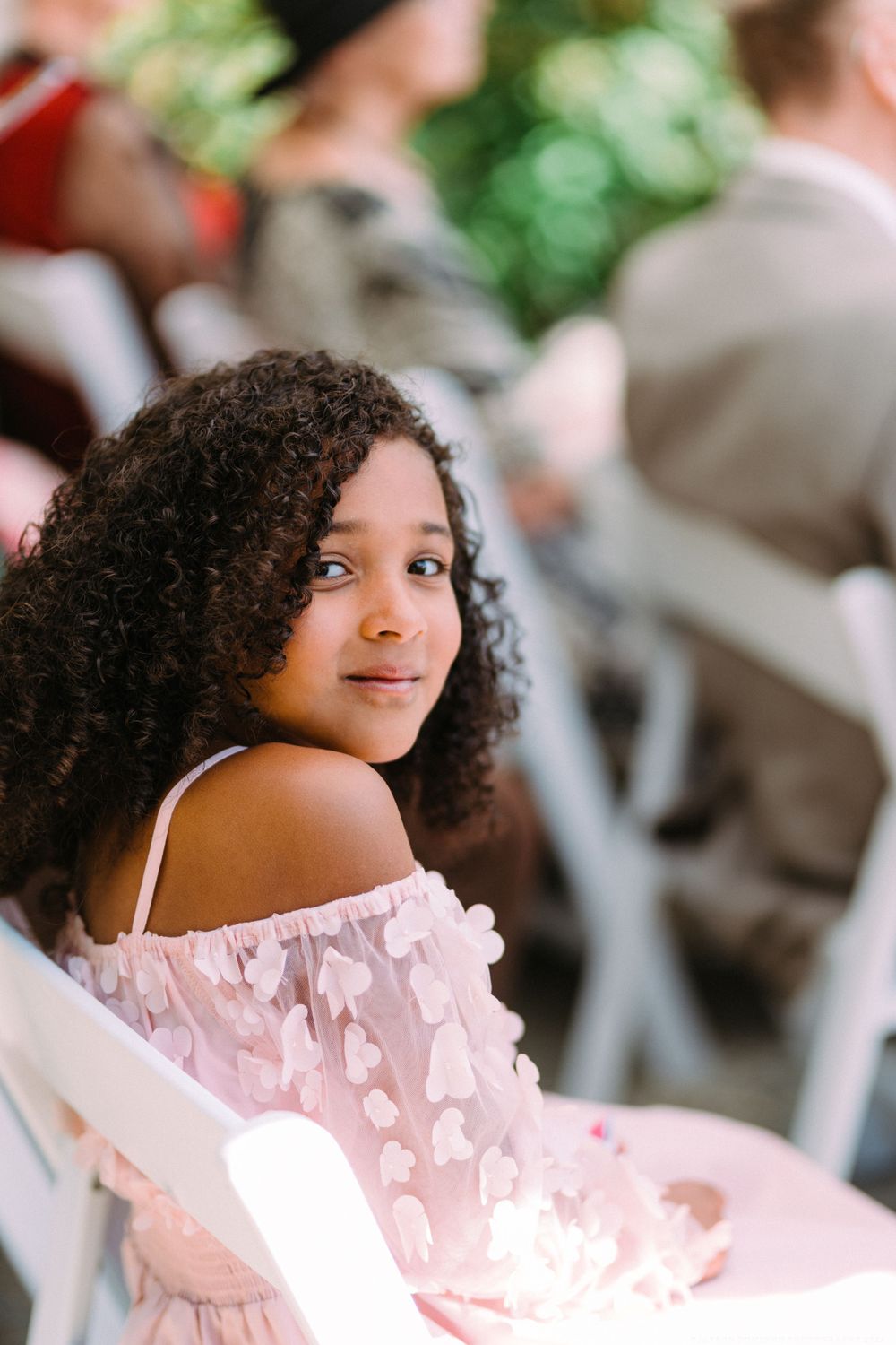 Wedding guest in pink off-shoulder dress sits in white chair at outdoor celebration.