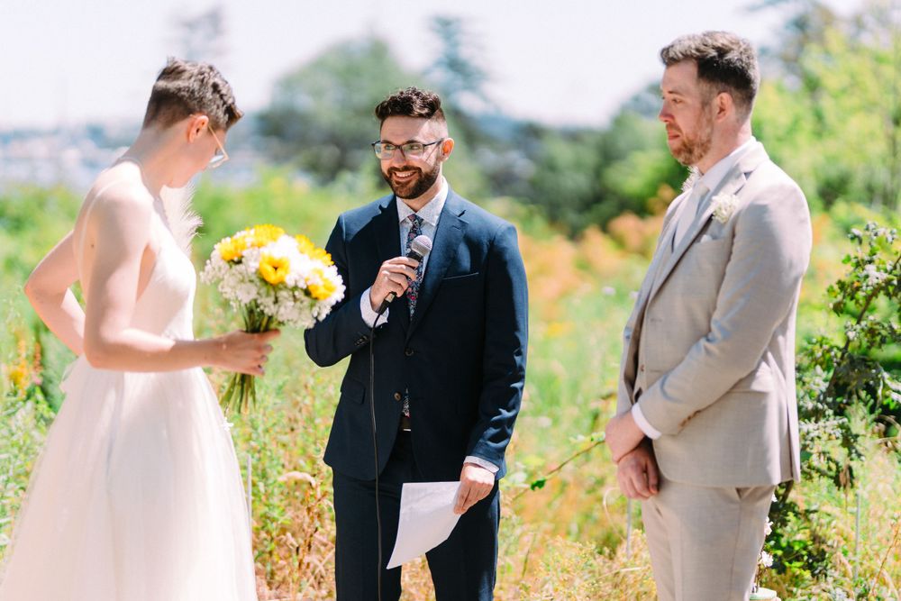 A wedding ceremony takes place outdoors with a couple and officiant surrounded by natural greenery and yellow flowers.