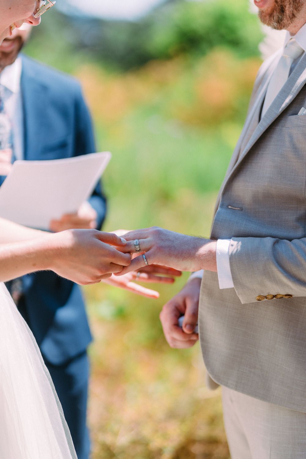 Two people exchange rings during an outdoor wedding ceremony with a light gray suit and white dress.