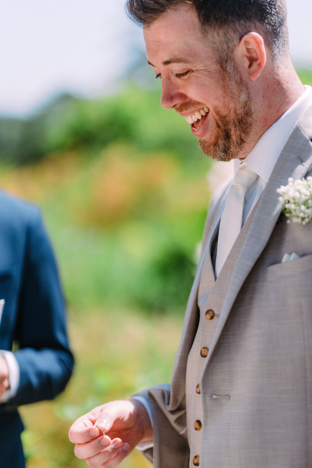 Person in light gray suit smiles joyfully during outdoor wedding ceremony.