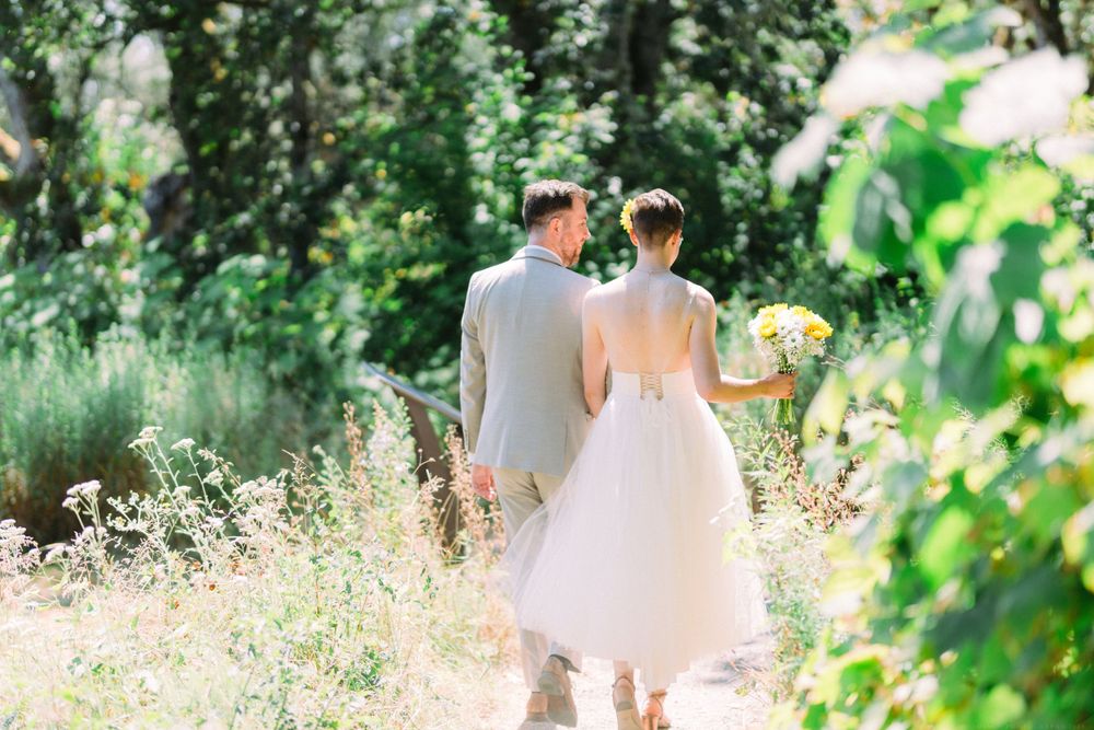 Couple walks through lush garden setting in wedding attire holding yellow sunflower bouquet.
