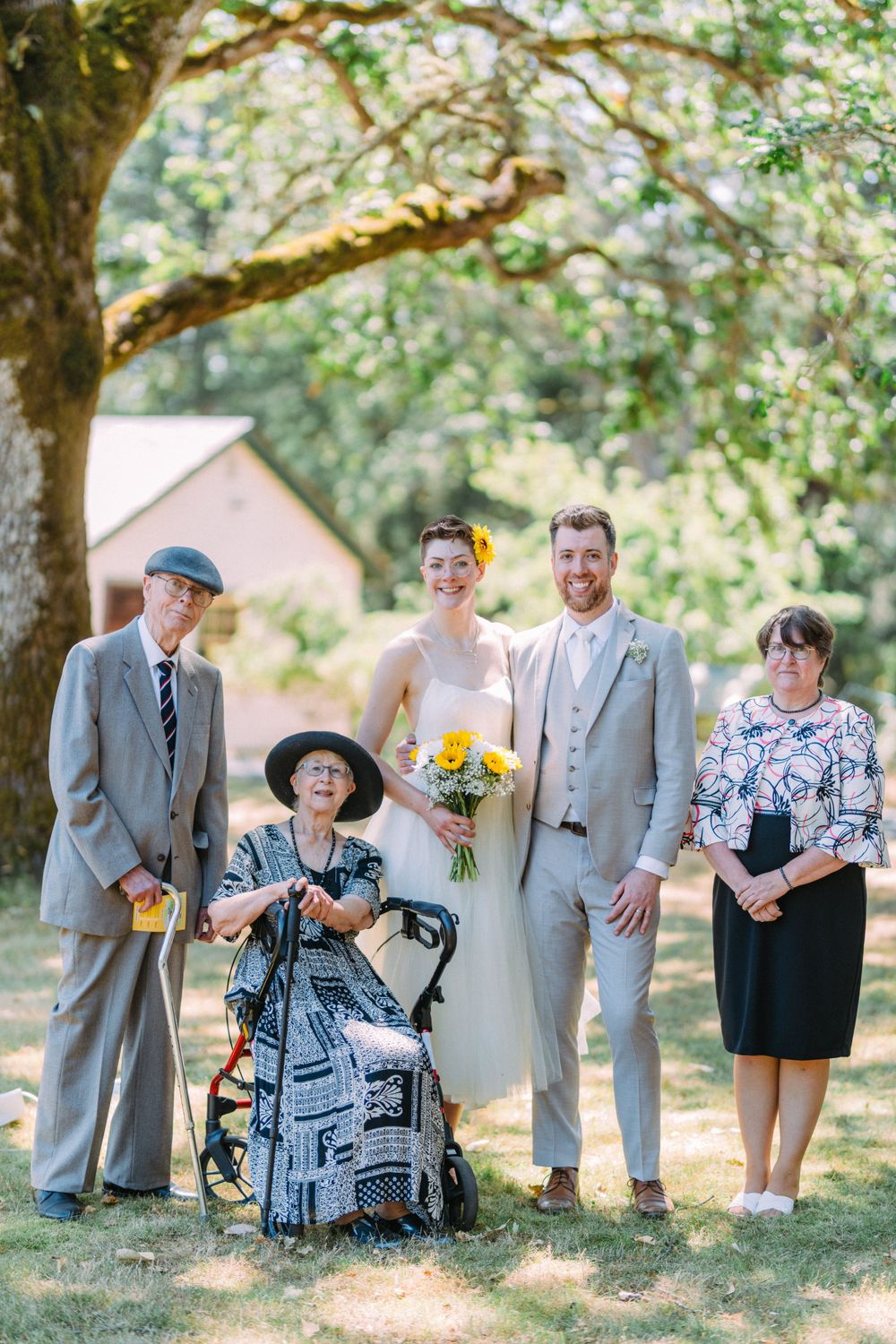 Wedding party poses outdoors under a tree with bride in white dress holding yellow flowers.