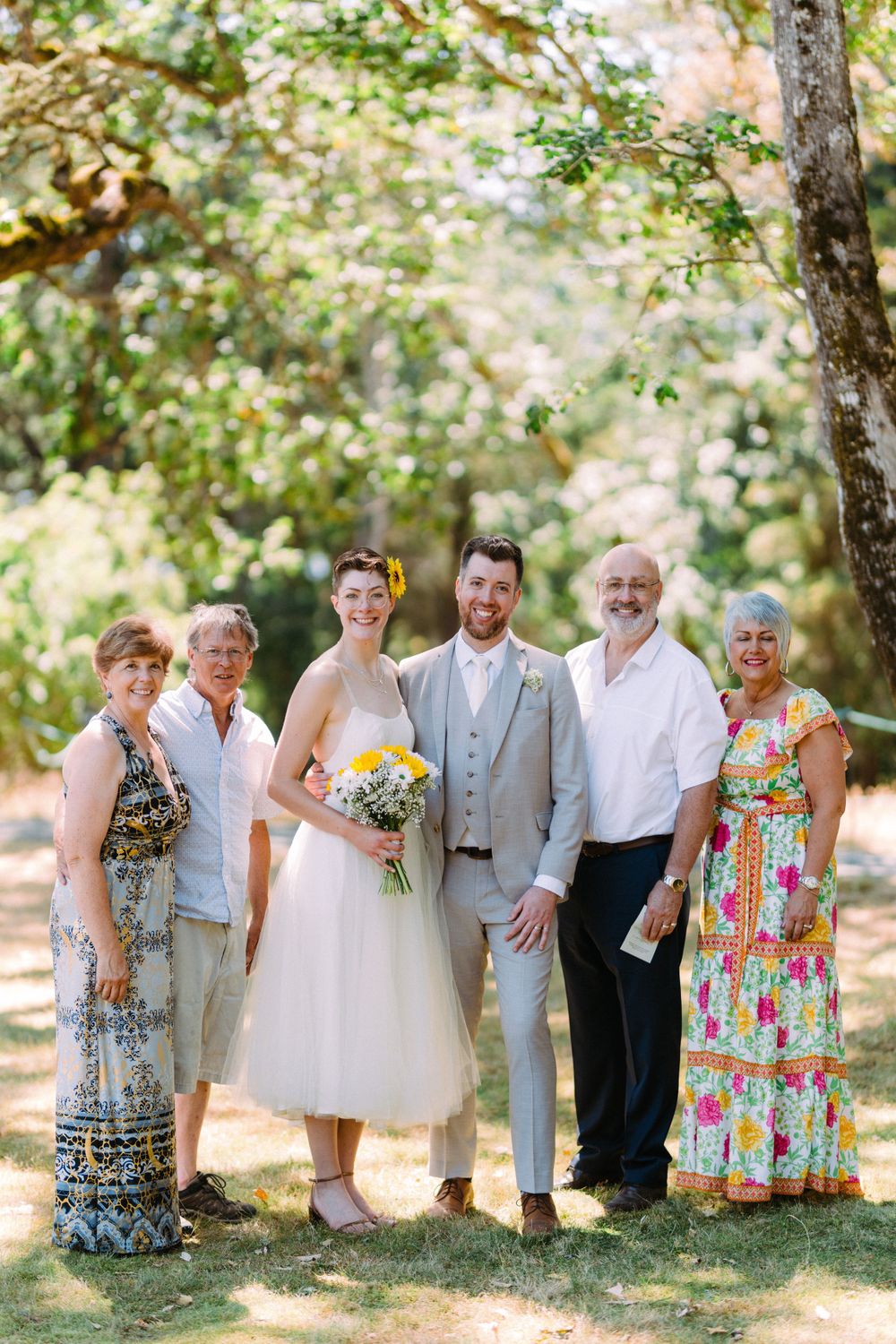 Wedding party gathers under trees for formal portrait on sunny day.