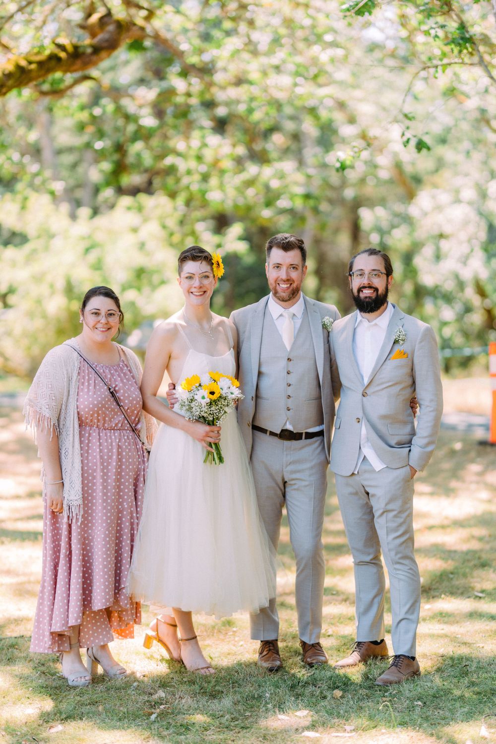 Wedding party of four poses under green trees with bride holding yellow bouquet.