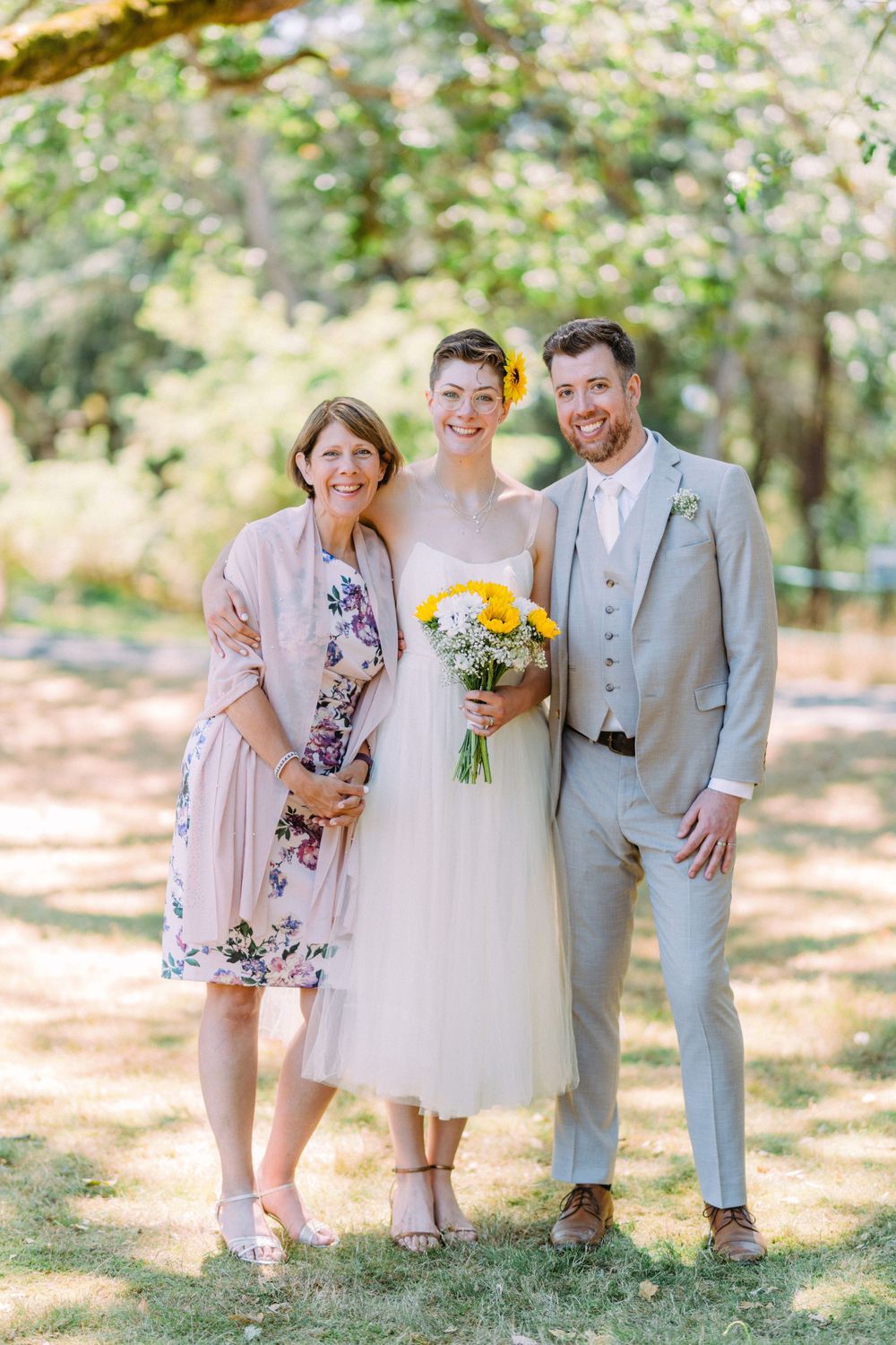 Outdoor wedding photo of three people with bride holding yellow flowers.