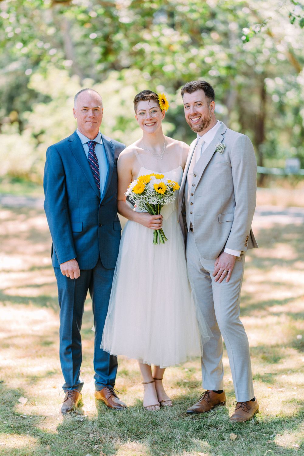 Wedding portrait of three people under sunny trees with bride in tea-length dress.