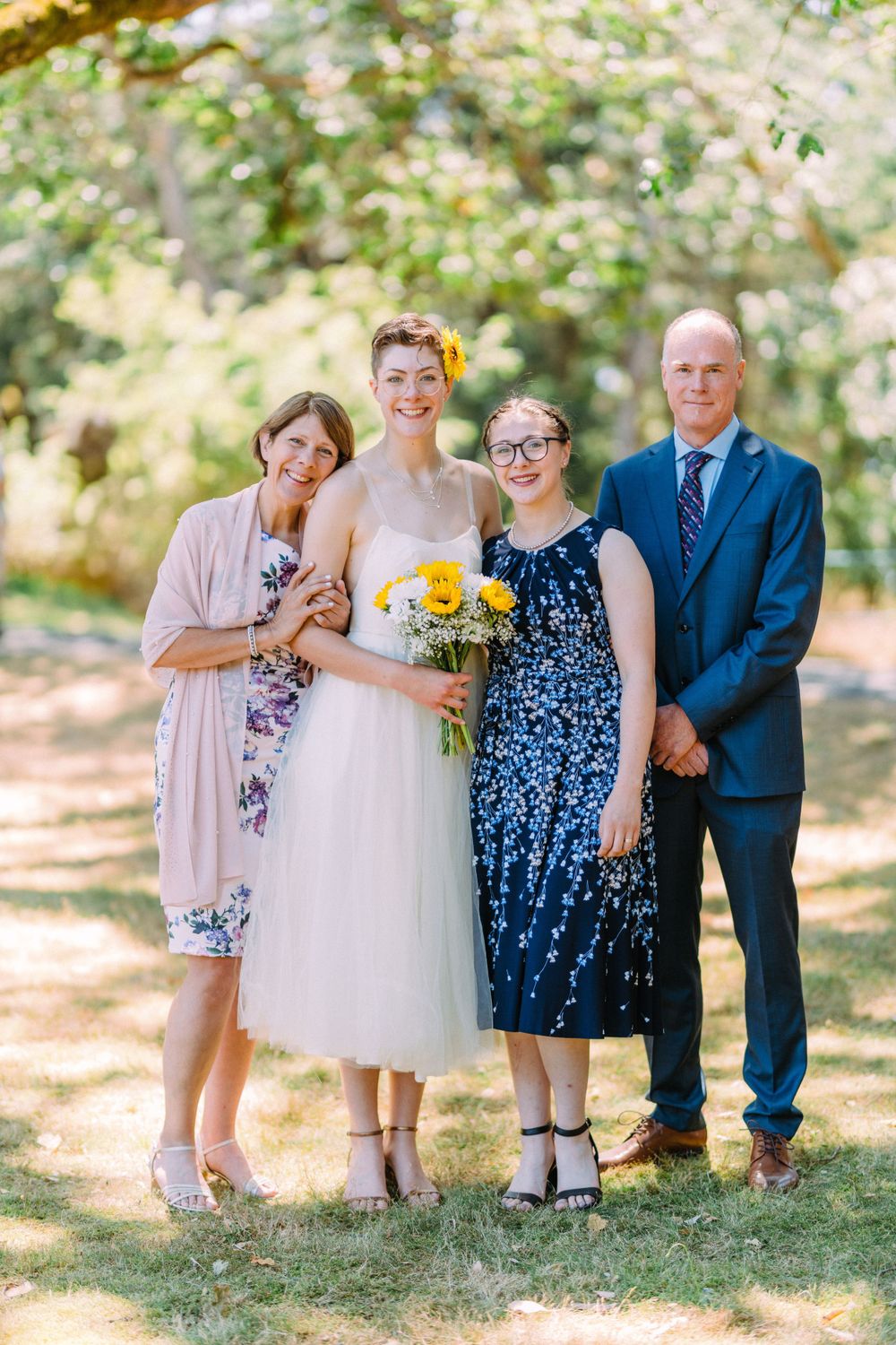 Wedding group photo with four people under trees with yellow bouquet and formal attire.