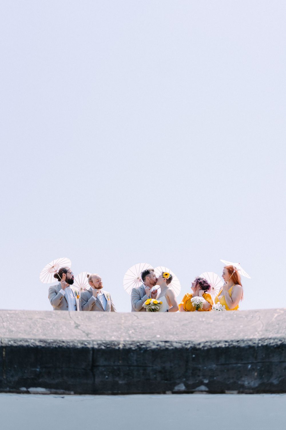 Wedding party sits casually on coastal wall while holding white parasols against bright sky.