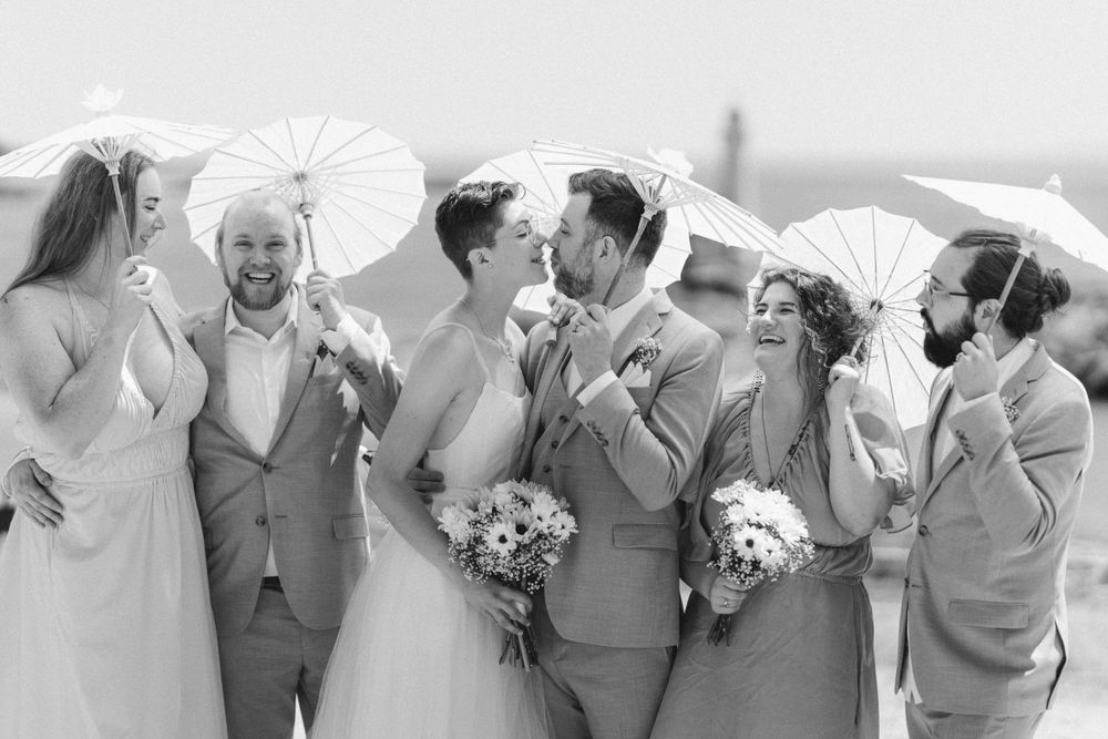 Wedding party poses together with white parasols in a black and white seaside portrait.