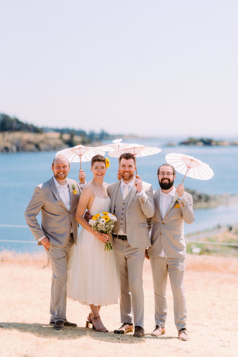 Wedding party in light suits and dresses pose with parasols on seaside cliff.