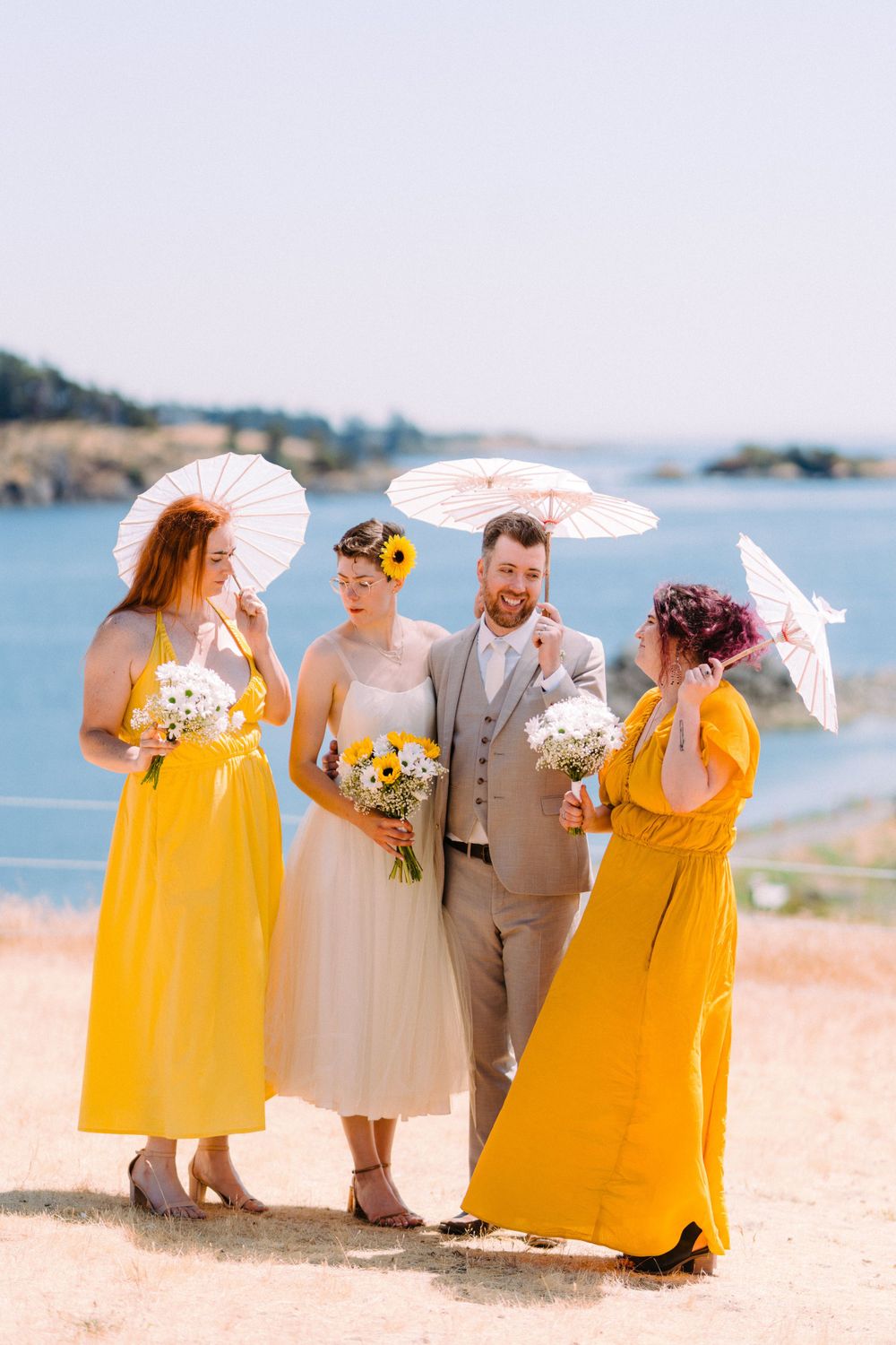 Wedding group in cream and yellow attire pose with parasols by ocean coastline.