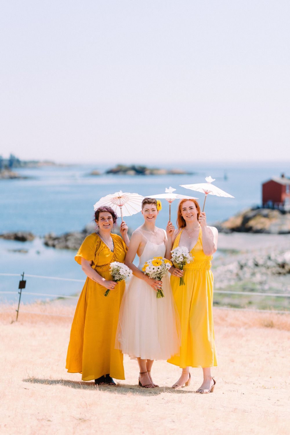 Three people in yellow and cream dresses pose with parasols against coastal backdrop.
