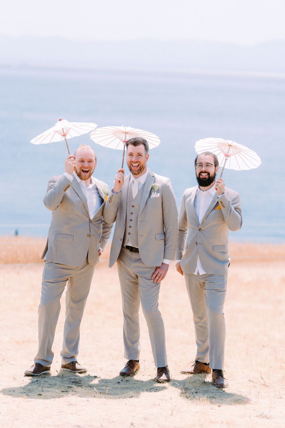 Three people in light gray suits share laughs while holding white parasols by the ocean.